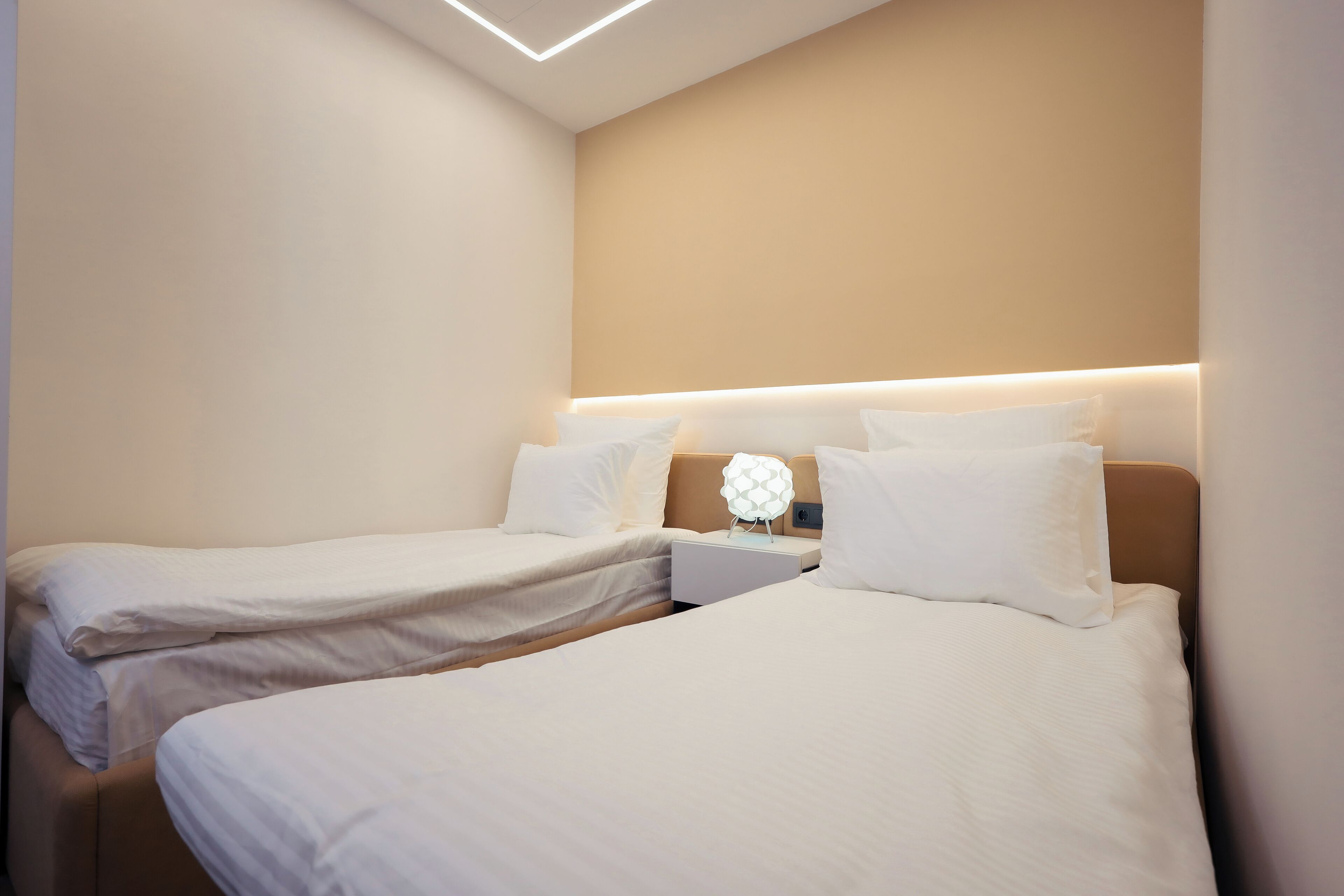 A hotel room with two single beds, a bedside table with a lamp, and a modern ceiling design.