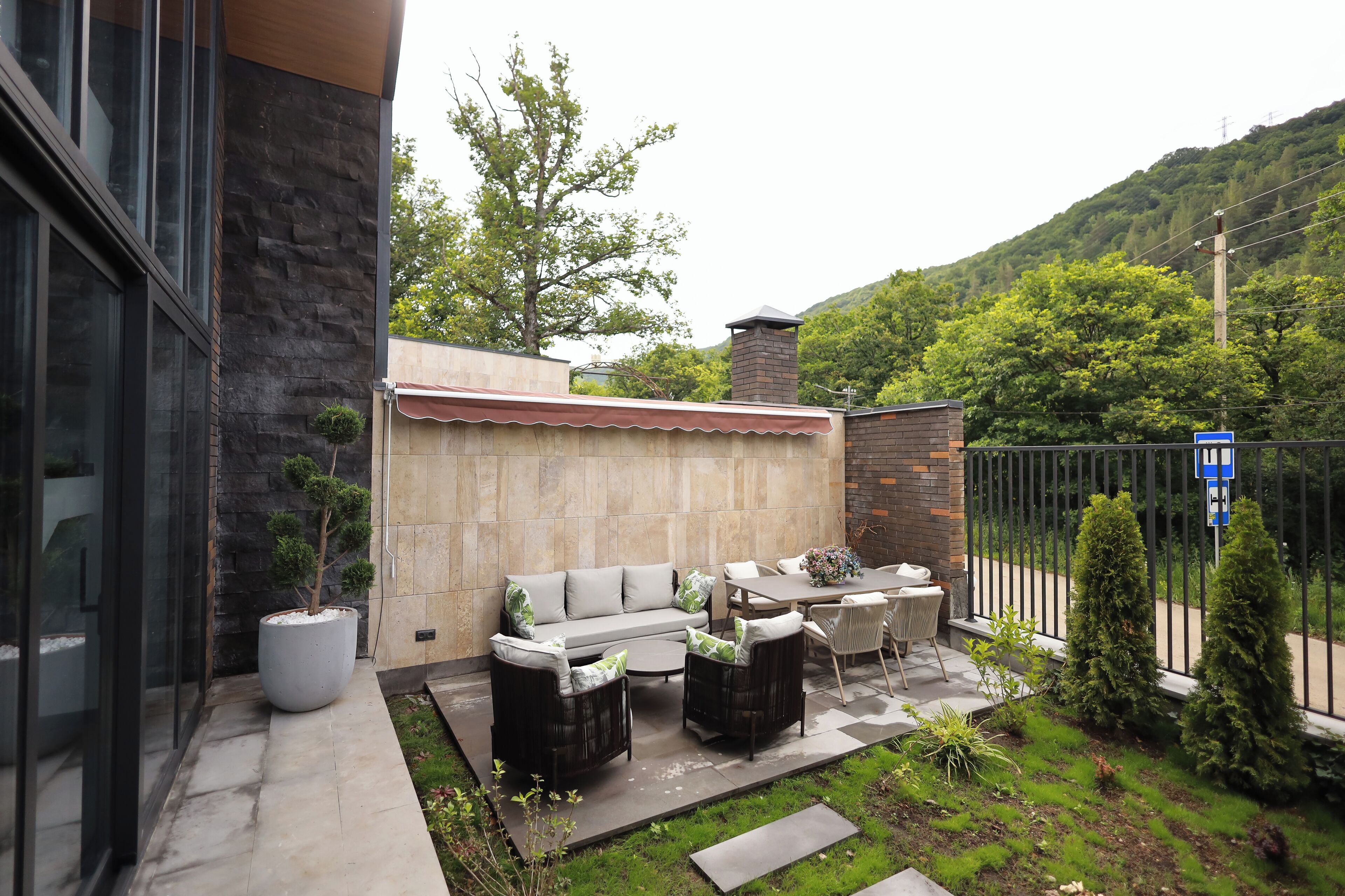 An outdoor patio with a sofa, chairs, and a table. There is a potted plant and a small tree. A fence and a gate are visible in the background.