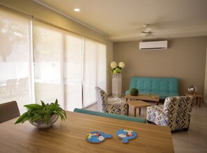 A modern living room with a teal sofa, patterned armchairs, a wooden coffee table, and a dining table with a plant and children's play mats. - Luxury 2 bedroom apartment (Cancún)