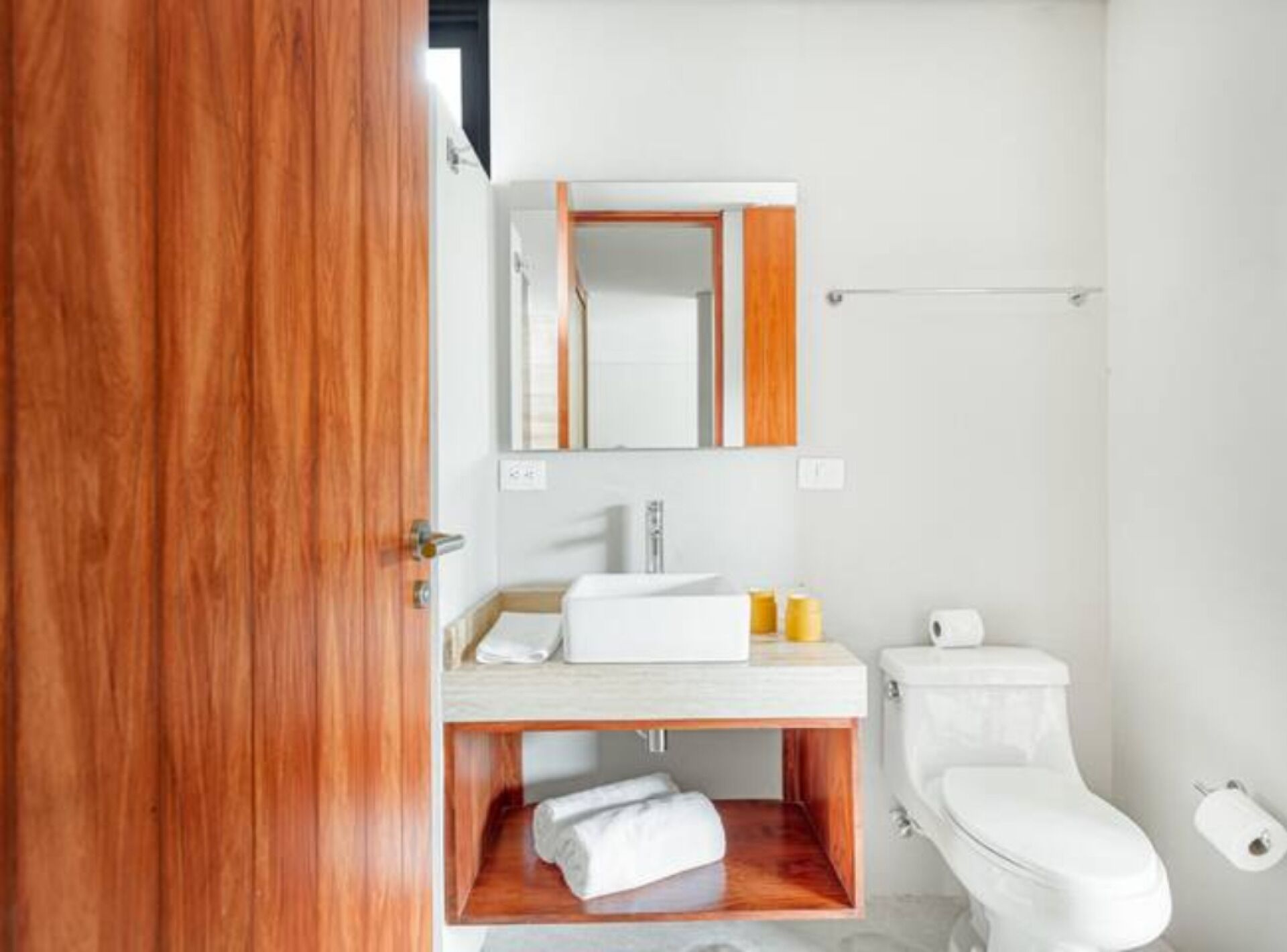 A modern bathroom with a wooden door, a white sink, a mirror, a toilet, and a towel shelf.