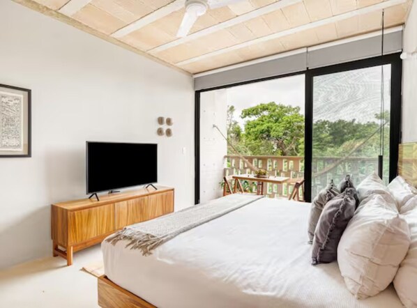 A bedroom with a large bed, a TV on a wooden cabinet, and a balcony with a view of trees.
