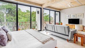 A spacious bedroom with a large bed, a sofa, a TV, and a balcony with a view of trees.