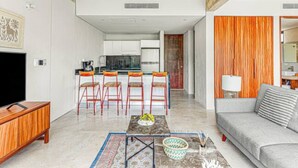 A modern living room with a sofa, a coffee table, and a TV. There is a kitchen area with a dining set and a wooden ceiling.
