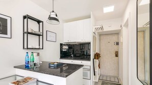 Una cocina moderna con armarios blancos, encimera negra y una lámpara colgante. Se observa una pequeña isla con superficie de vidrio y una revista encima.