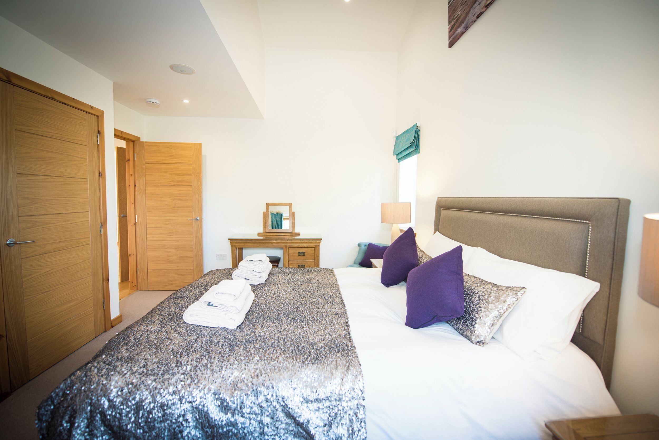 A bedroom with a large bed, wooden wardrobe, a small table with a mirror, and a nightstand with a lamp.
