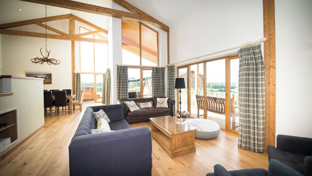 A spacious living room with a high ceiling, wooden beams, a large sofa, a coffee table, and a view through a sliding glass door to an outdoor deck. - Atlas (Nairn)