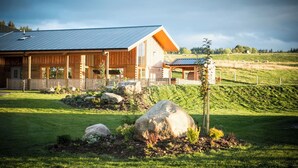 A large wooden lodge with a metal roof, a covered porch, and a fenced yard with large rocks and a small tree. - Atlas (Nairn)
