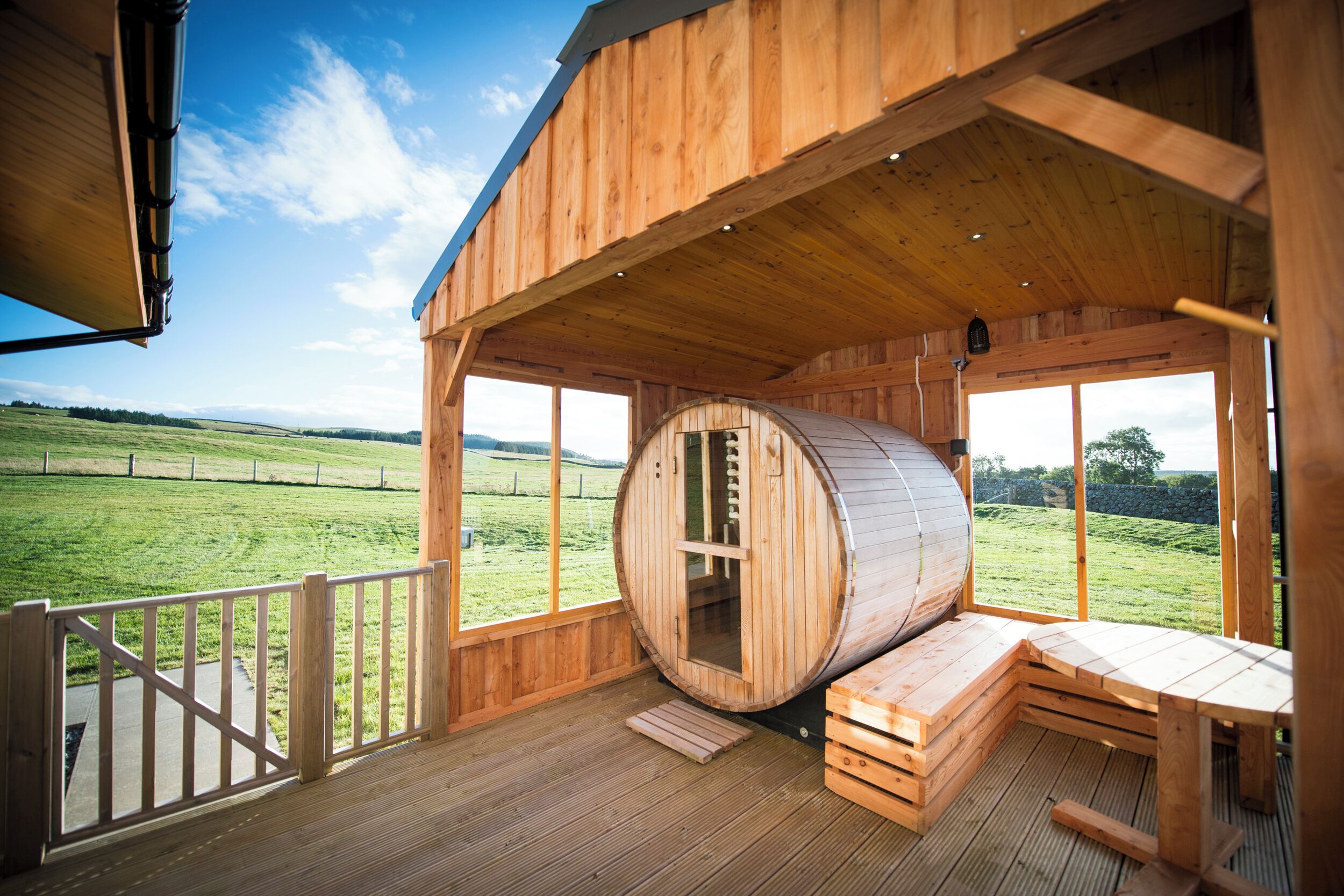 A wooden cabin with a sauna, large windows, and a deck overlooking a green landscape.