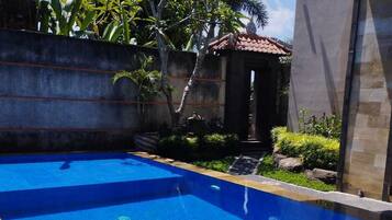 A rectangular swimming pool with clear blue water, surrounded by a concrete wall and a stone building with a tiled roof.