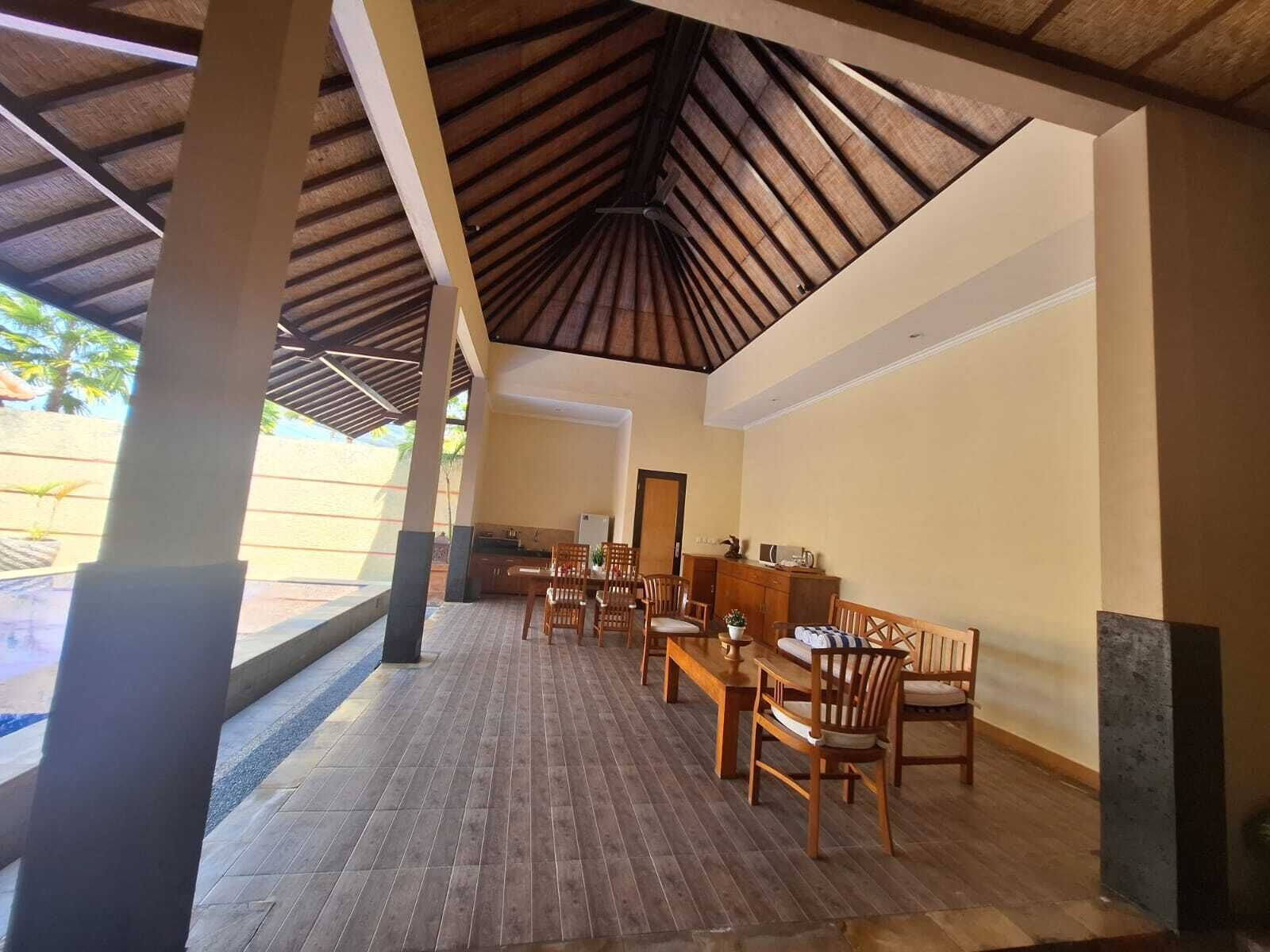 A covered outdoor dining area with wooden tables and chairs, a kitchen in the background, and a view of the outdoors.