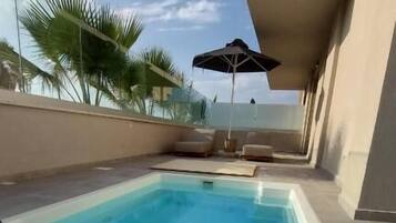 A rectangular swimming pool with a white edge, surrounded by a tiled deck. There are lounge chairs and a single umbrella. Palm trees are visible in the background.