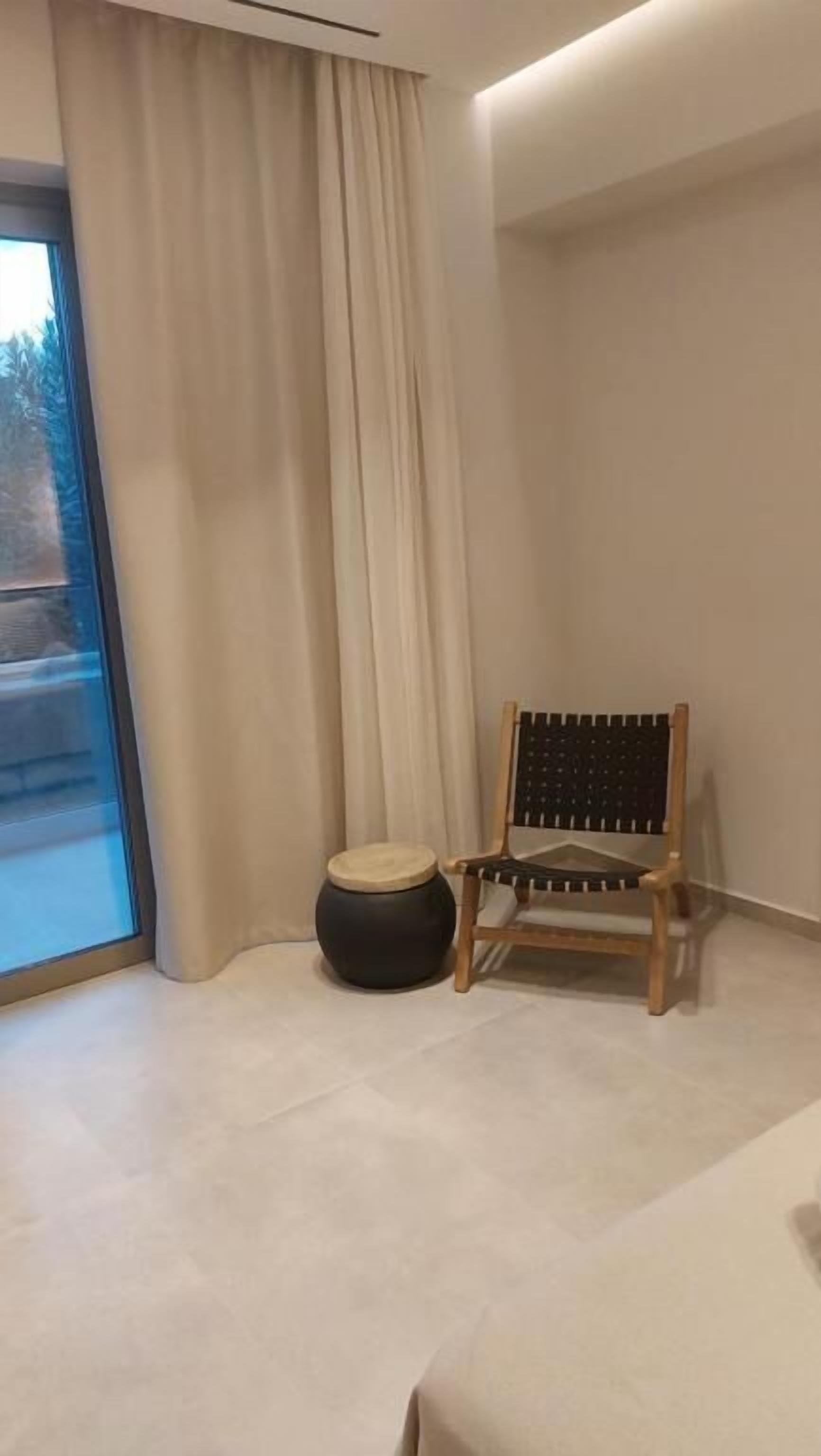 A corner of a room with a wooden chair, a round ottoman, and a large window with curtains.