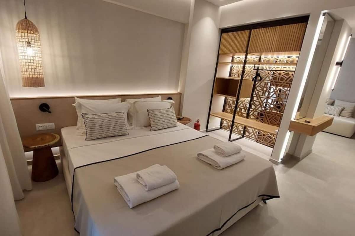Ein modernes Hotelzimmer mit einem großen Bett, Nachttischen, einer Hängelampe und Blick auf eine Holzkonstruktion außerhalb.