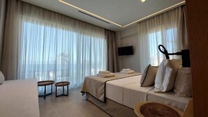 A modern hotel room with a bed, two small side tables, a flat-screen TV mounted on the wall, and large windows with sheer curtains. - Niriides Palace (Hersonissos)
