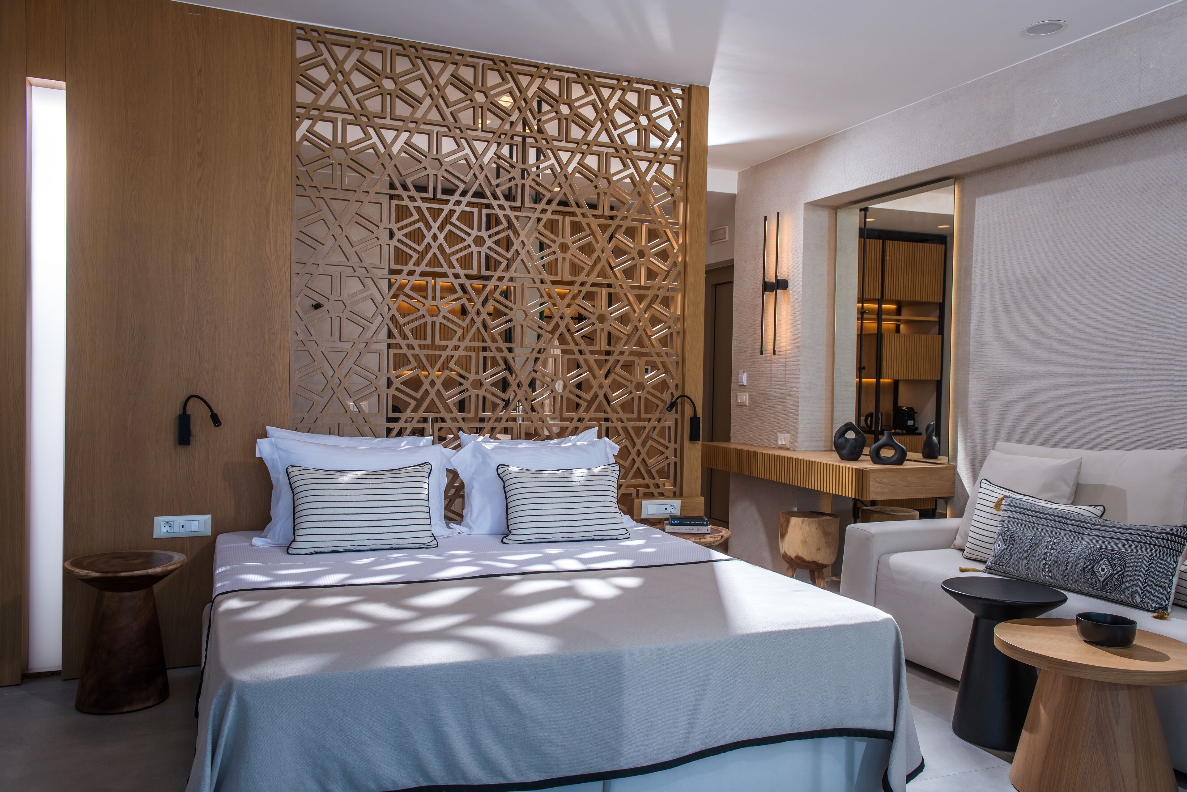 A modern hotel room with a large bed, a wooden partition, a sitting area with a sofa and a small table, and a mirror above the sofa.