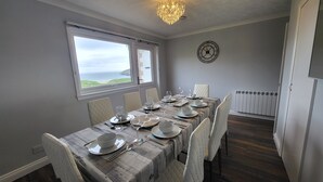Comedor con una mesa dispuesta para seis personas, vista al océano a través de una ventana, un reloj en la pared y un radiador en la esquina.