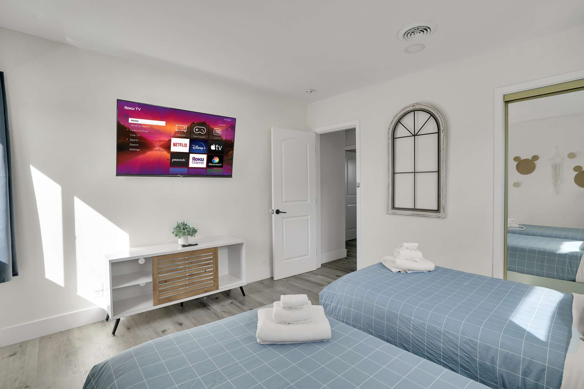 Une chambre d’hôtel moderne dotée d’un grand téléviseur à écran plat affichant l’interface Roku, d’une armoire en bois avec une plante, de deux lits avec une literie bleue et d’une petite salle de bain avec un miroir et un porte-serviettes.