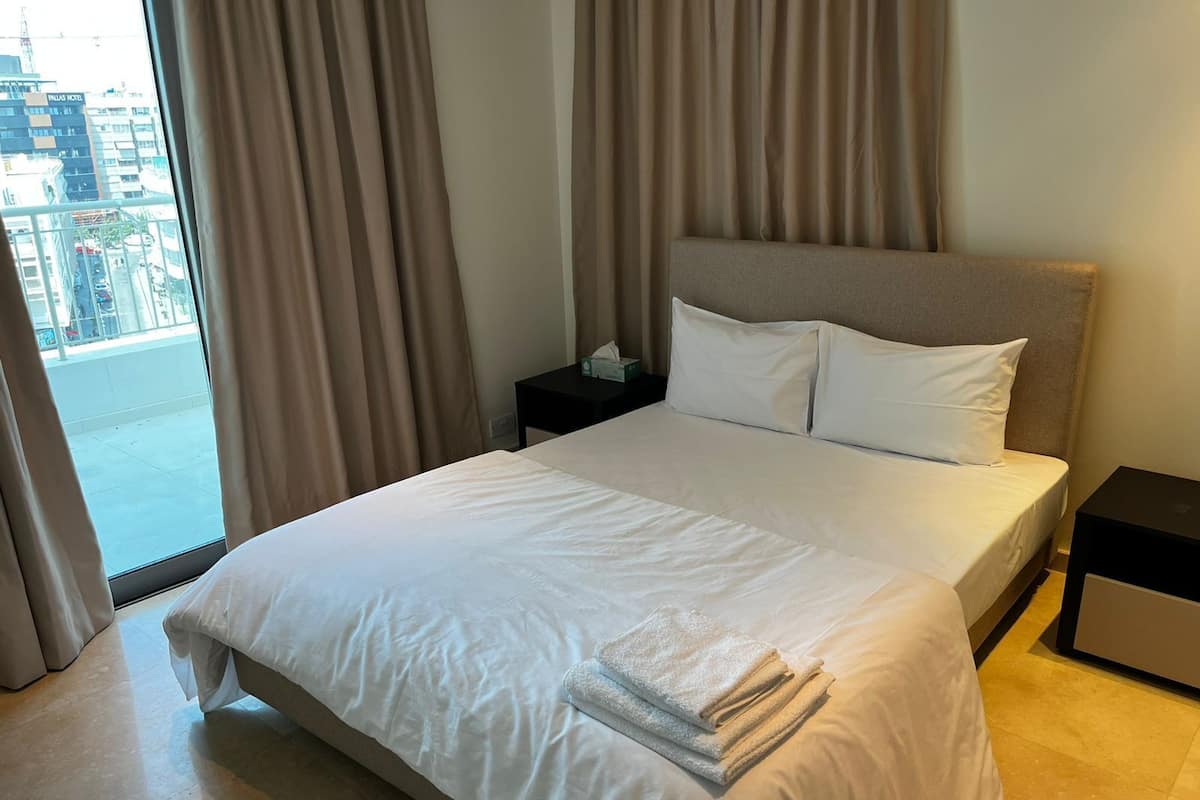 A hotel room with a large bed, a nightstand, a window with curtains, and a view of the city.