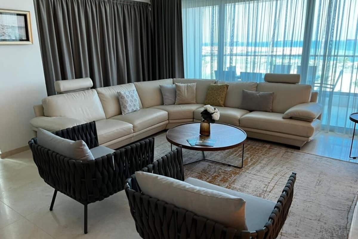 A modern living room with a sectional sofa, two armchairs, a coffee table, and a view of the sea through large windows.