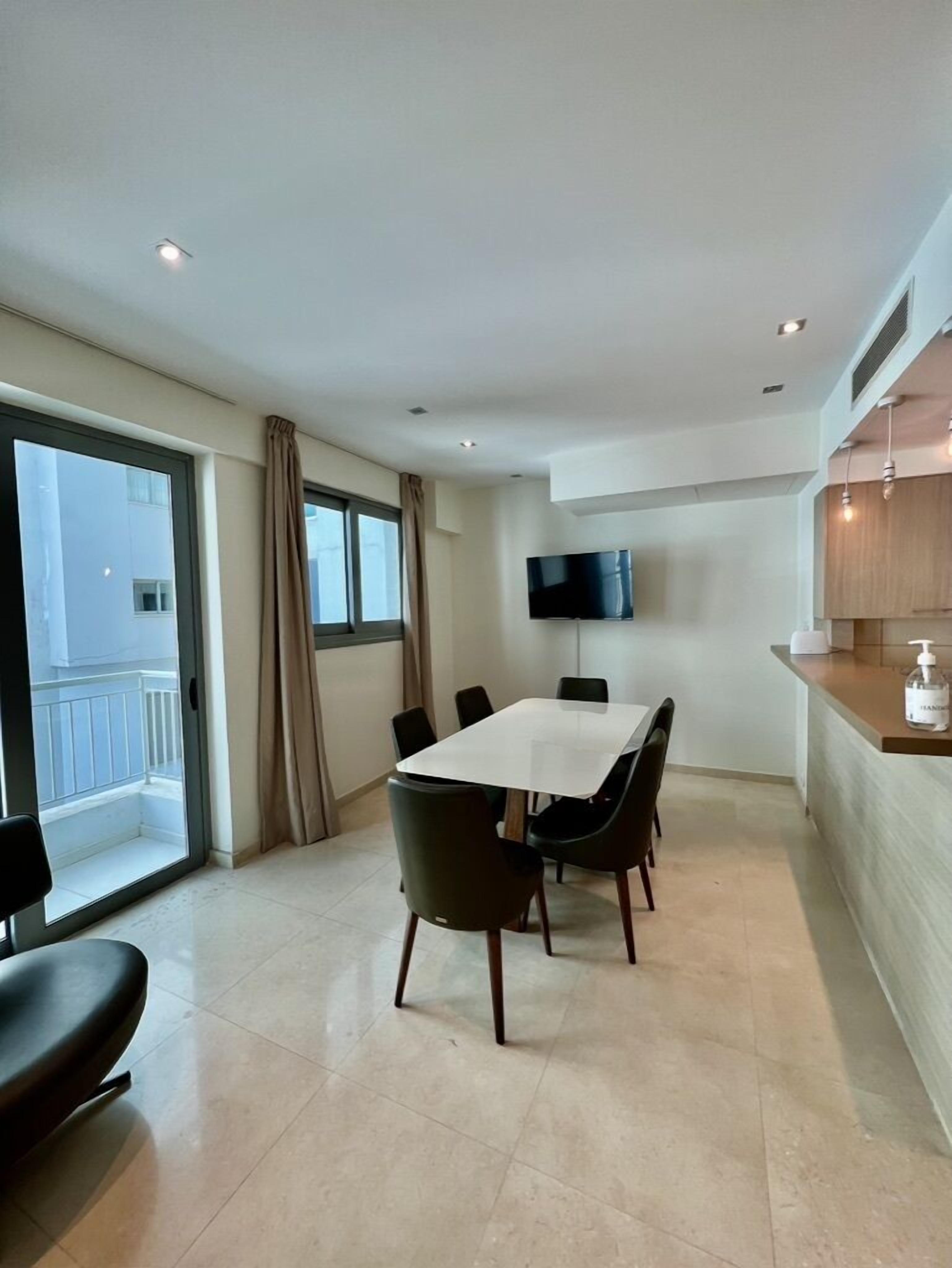 Un espace repas moderne avec une table et des chaises, un téléviseur fixé au mur et un comptoir de cuisine équipé d’un distributeur de savon.