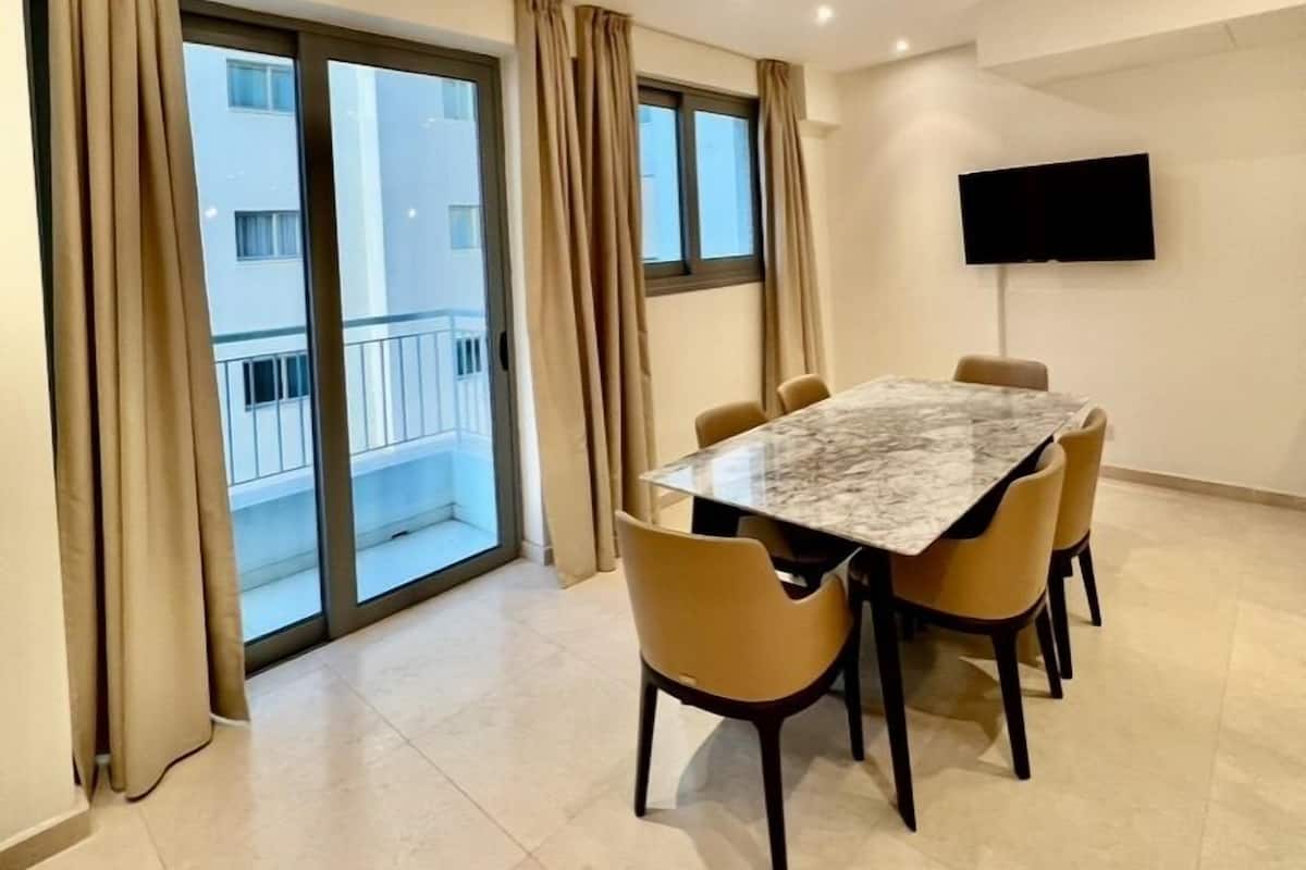 A modern dining area with a marble table and chairs, large windows with curtains, and a flat-screen TV mounted on the wall.