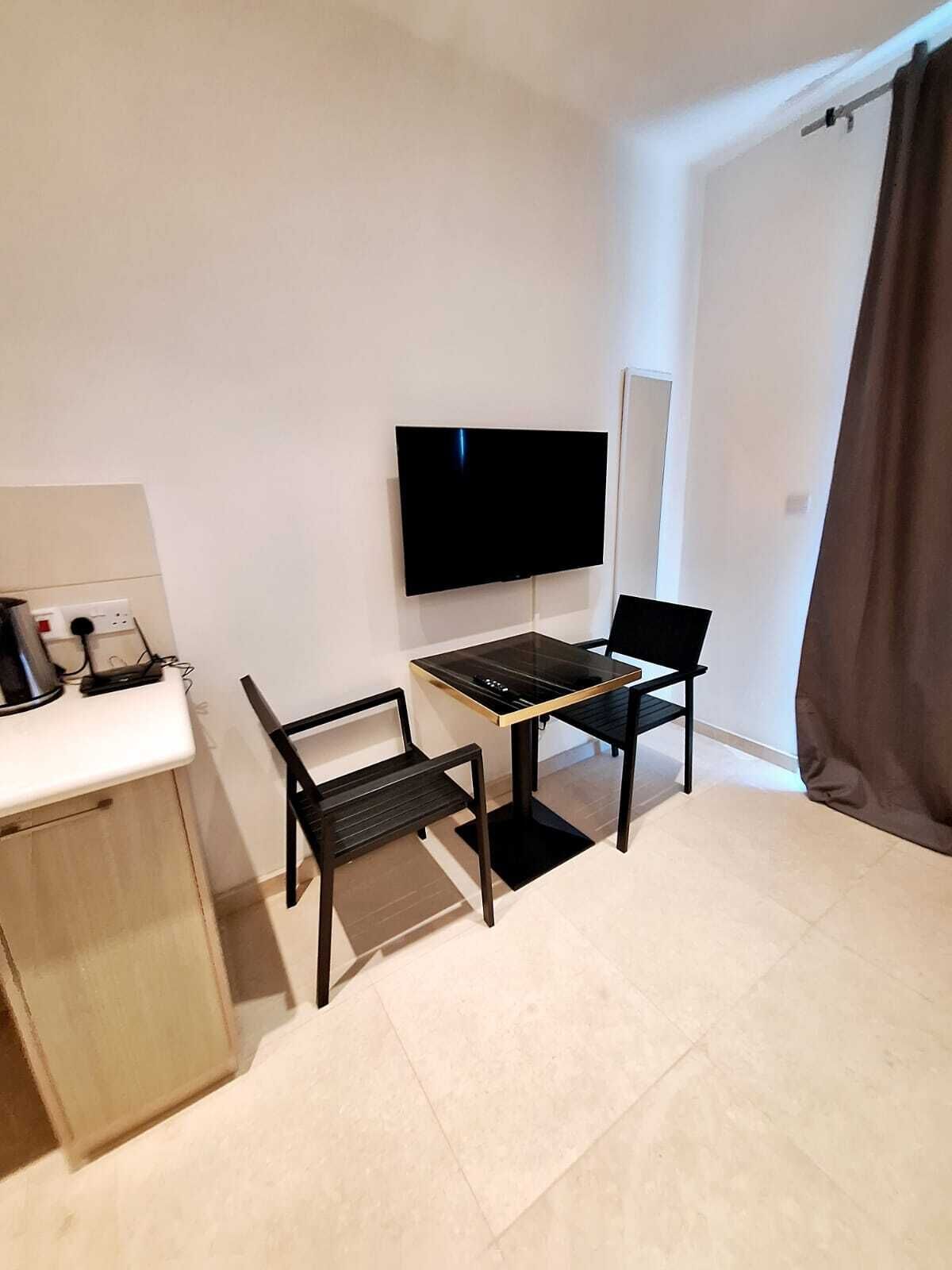 Una habitación pequeña y moderna con televisor de pantalla plana en la pared, una kitchenette compacta con cafetera y una sola silla.