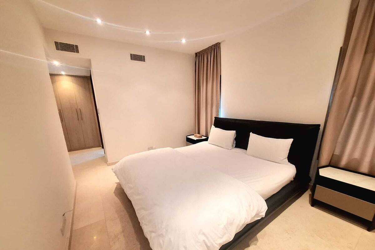 A modern hotel bedroom with a large bed, a bedside table, and a wardrobe.