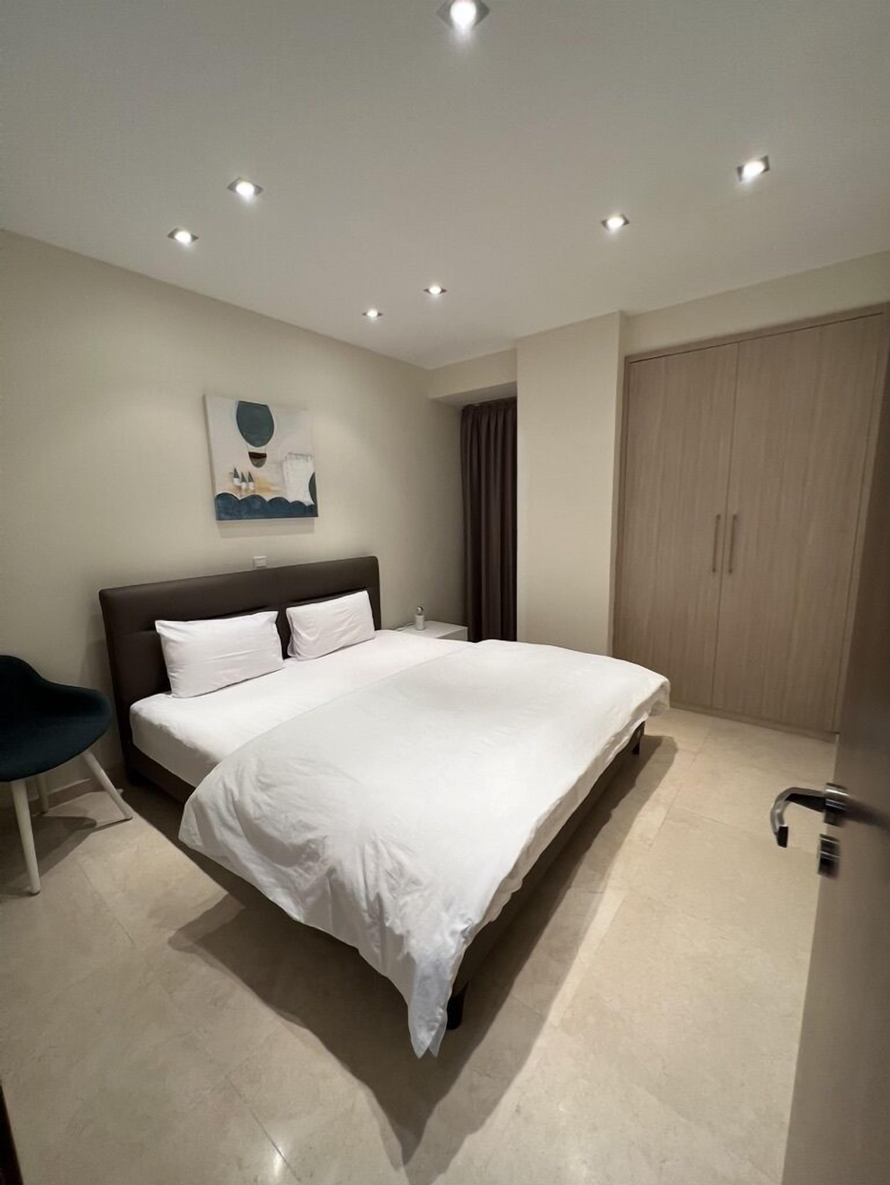 A modern bedroom with a large bed, a nightstand, a chair, a wardrobe, and recessed ceiling lights.