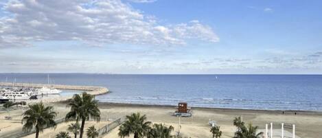 A beach view from a balcony with palm trees, a pier, and boats in the distance.