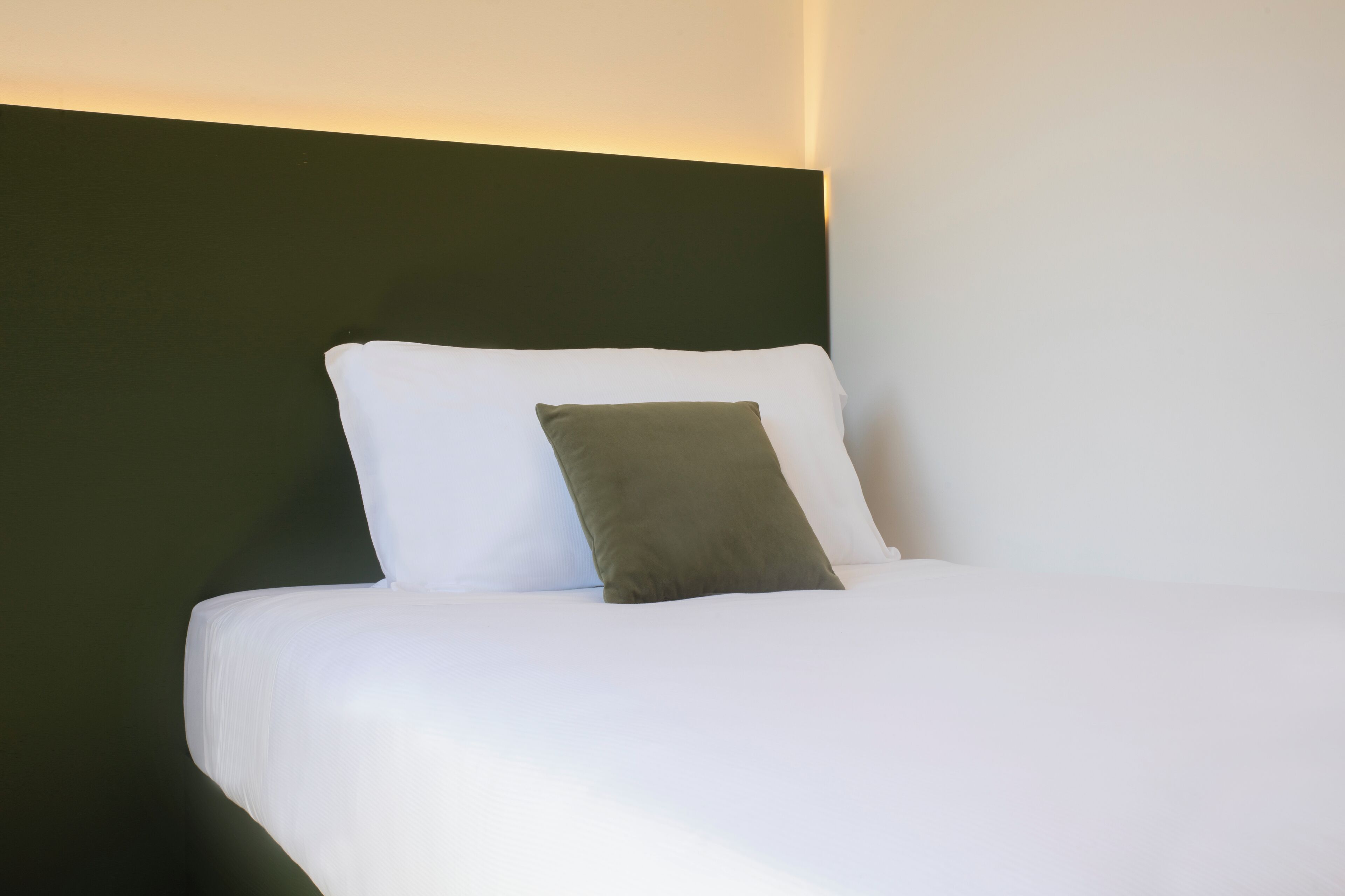 A neatly made bed with white and green pillows, a dark headboard, and a light wall.