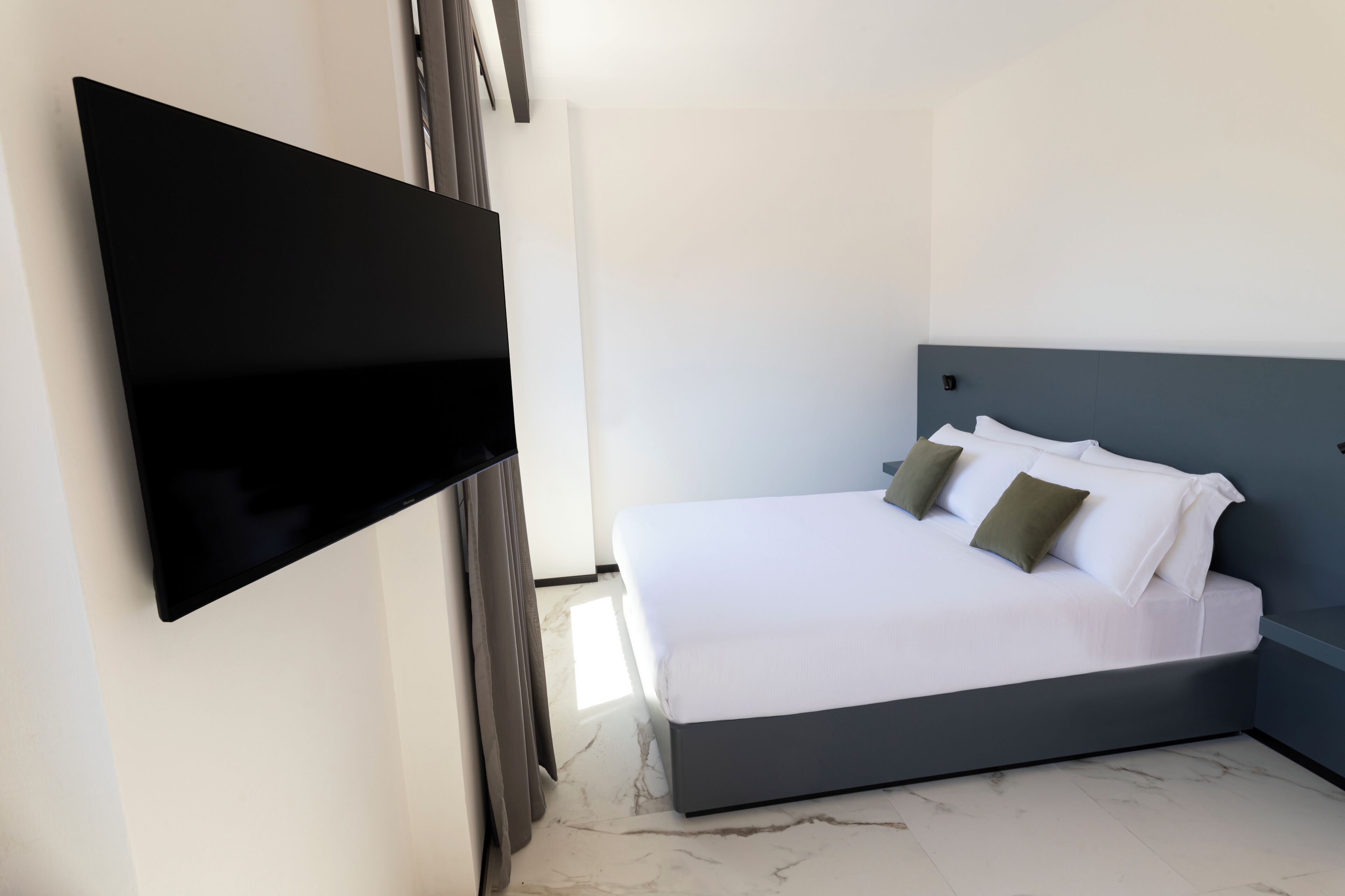 A modern hotel room with a large bed, a flat-screen TV mounted on the wall, and a marble floor.