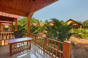 Una terraza de madera con una mesa y sillas, rodeada por una barandilla de madera, con vistas a un jardín tropical con palmeras y un edificio con techo rojo.