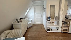Ein schmales Dachzimmer mit schräg stehender Decke, einer weißen Tür, einem kleinen weißen Schreibtisch, einem Stuhl und einem Bücherregal.