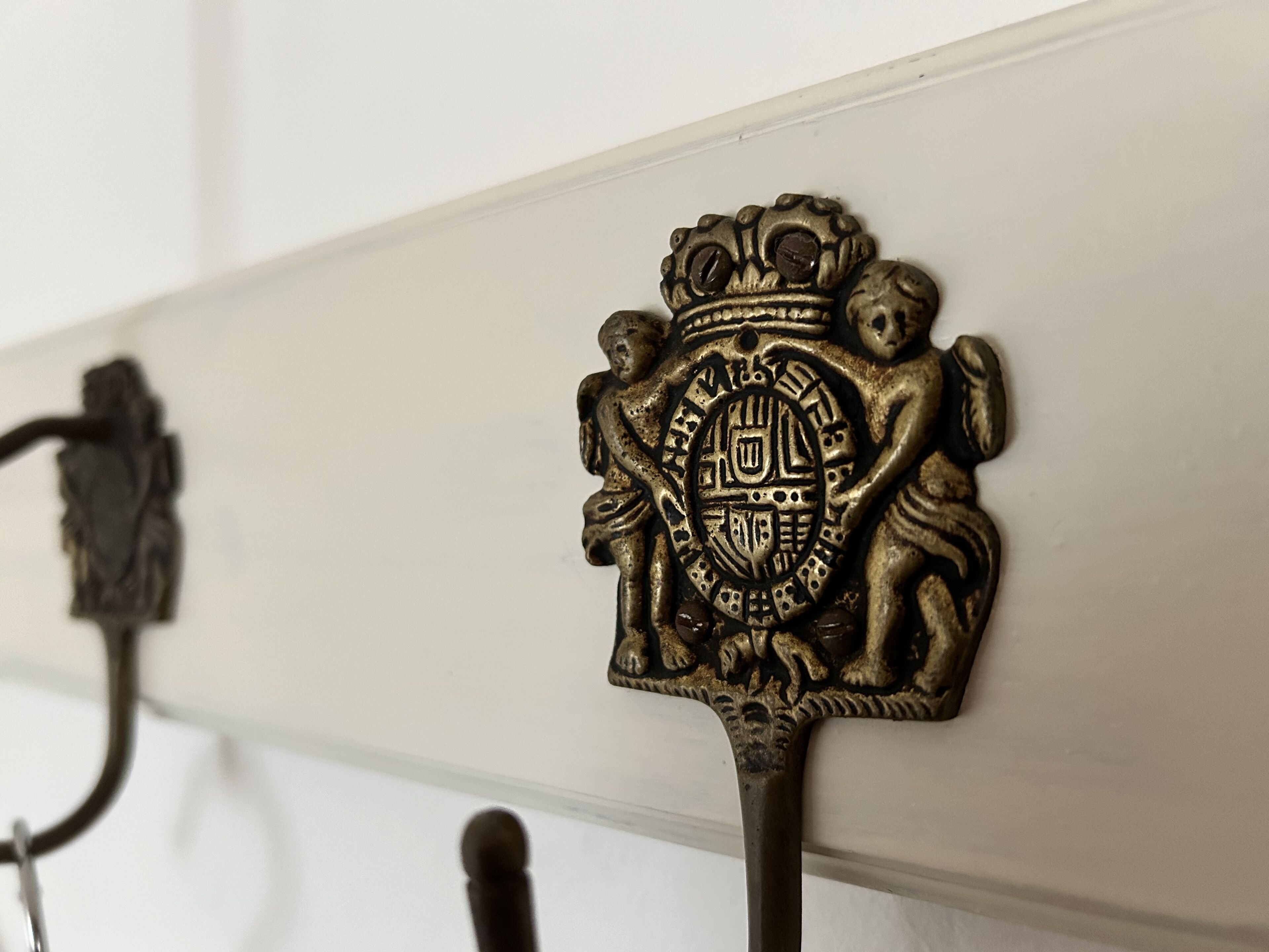 A close-up of a brass coat hook with an ornate emblem, mounted on a white wall.