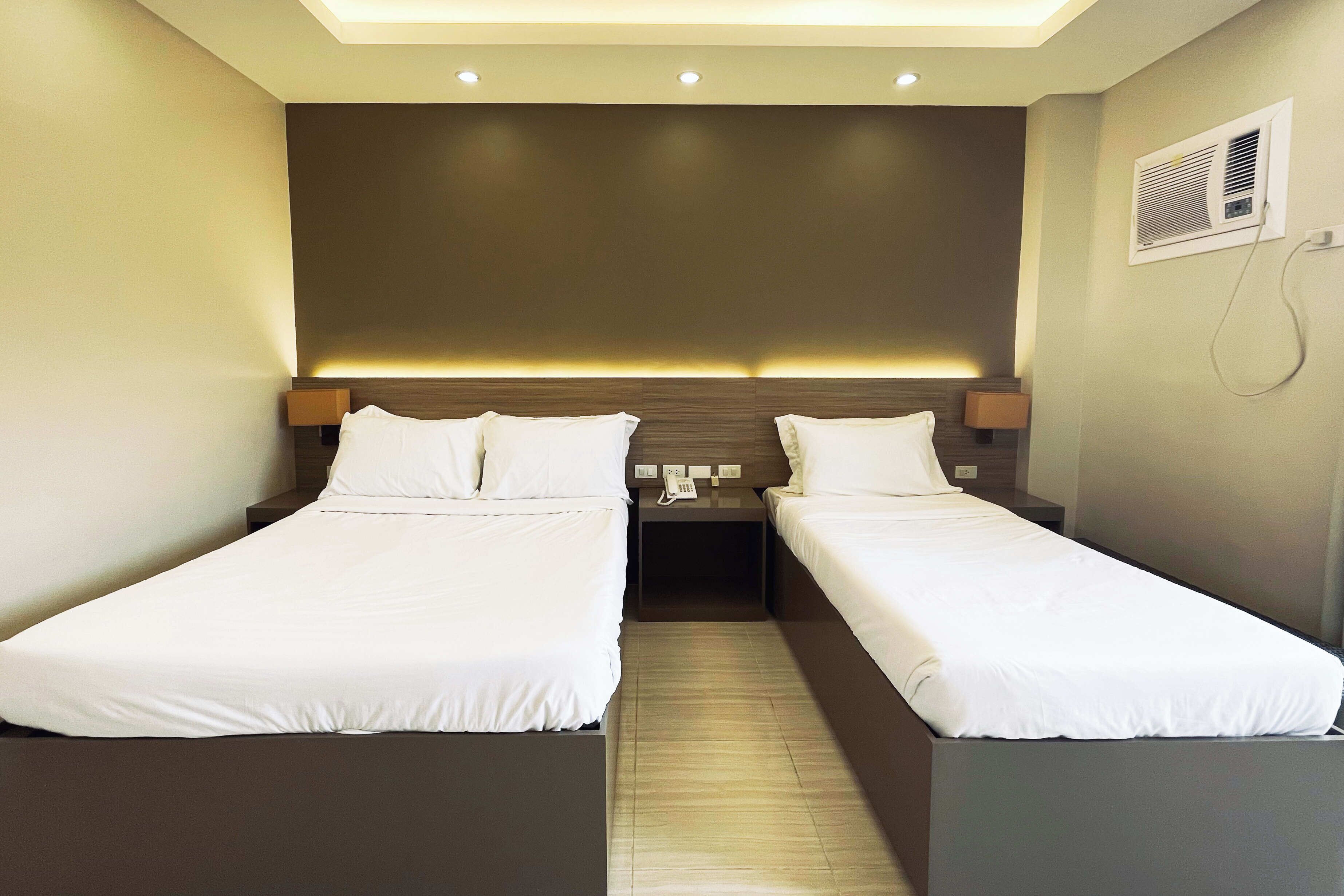 A hotel room with two single beds, a nightstand with a phone, and a wall-mounted air conditioning unit.