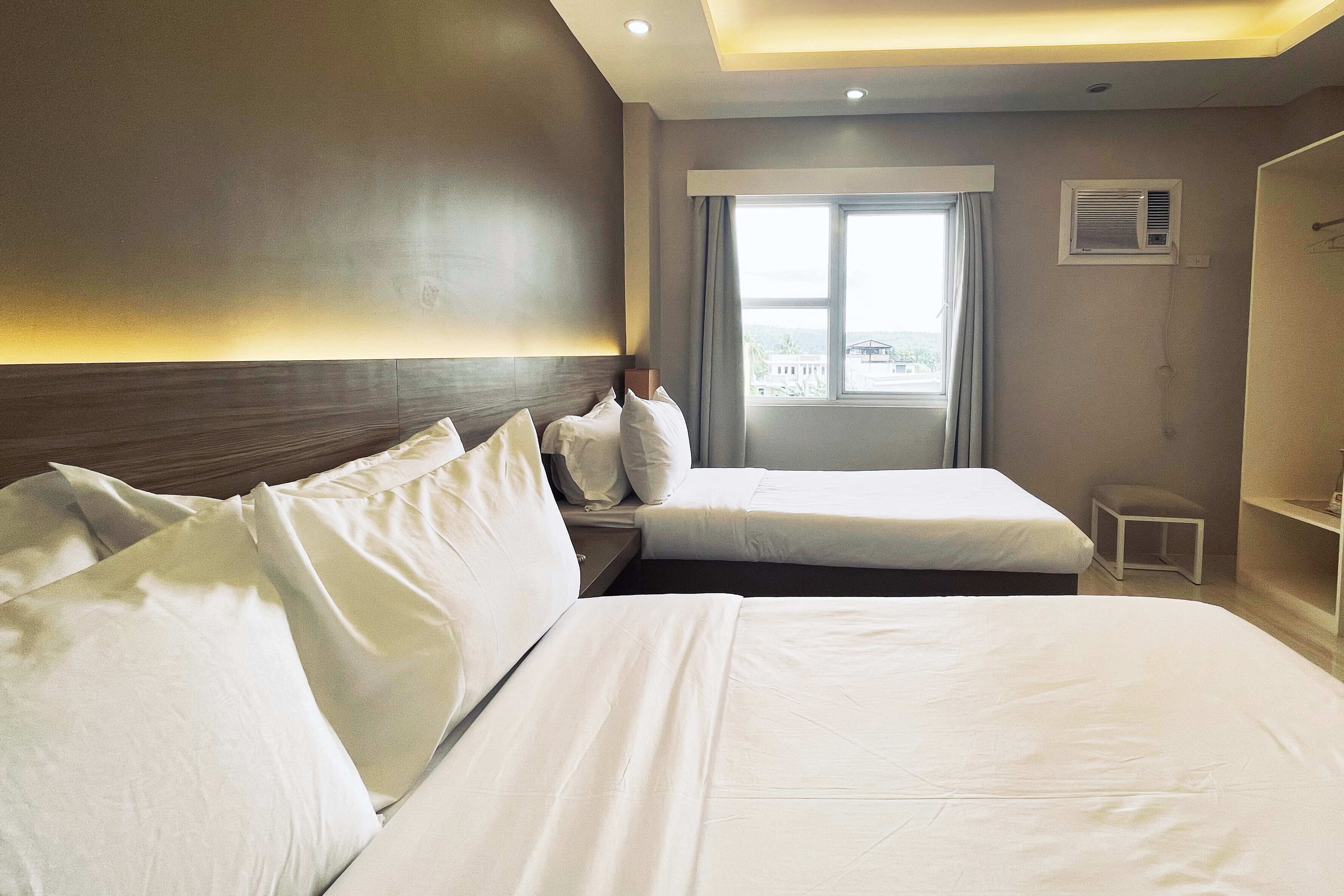 A hotel room with two beds, a window with curtains, an air conditioning unit, and a small white stool.