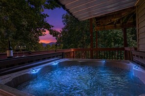 A hot tub with a wooden deck and a view of trees and mountains at sunset.