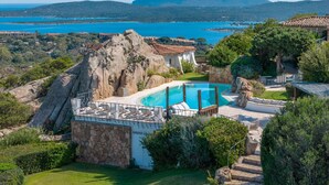 A swimming pool surrounded by stone walls and greenery, with a view of the sea and mountains in the background.