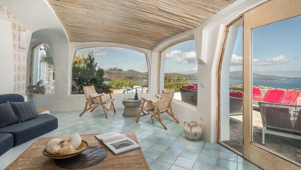 A spacious room with a large window offering a scenic view of the sea and mountains. There are two wicker chairs and a wooden table with a book and decorative items.