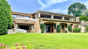 A two-story house with a covered patio, a lawn, and flowering plants in the foreground. - Villa in Porto Rotondo with 7 bedrooms sleeps 14 (Porto Rotondo)
