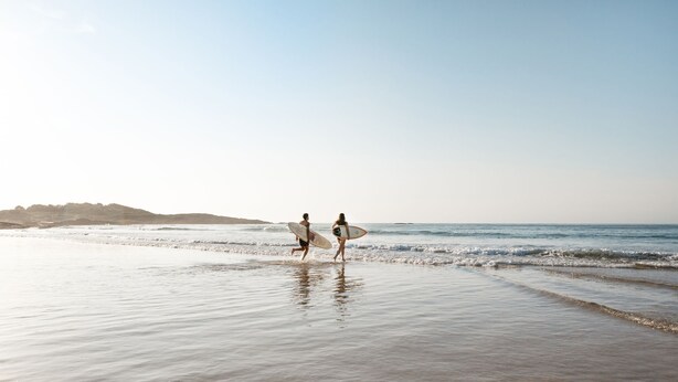 Escena de playa con dos personas corriendo con tablas de surf.