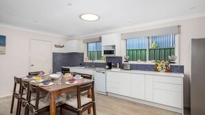 Una cocina funcional con armarios blancos, microondas y lavamanos. Hay una mesa de comedor con sillas y un ventanal con persianas verdes.