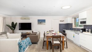 Una sala de estar moderna con un televisor de pantalla plana, una mesa de comedor para cuatro personas y una cocina con armarios blancos y un microondas.
