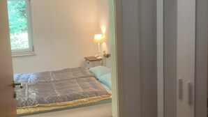 A bedroom with a bed, a nightstand, a lamp, a wardrobe, and a window with a view of greenery. - Immensee II Apartment with lake view and located at the forest edge (Ronshausen)