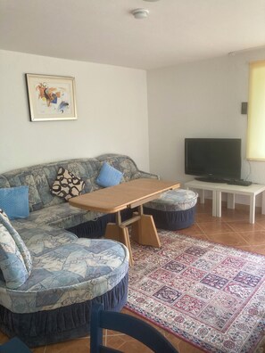 Un salon comprenant un canapé, un pouf, une table basse en bois et un téléviseur posé sur un meuble.