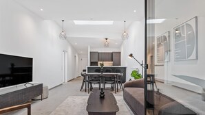 Een moderne woonkamer met een flatscreen-televisie, een donker houten salontafel en een bank. Er is een eethoek met een tafel en stoelen, en een keuken op de achtergrond.