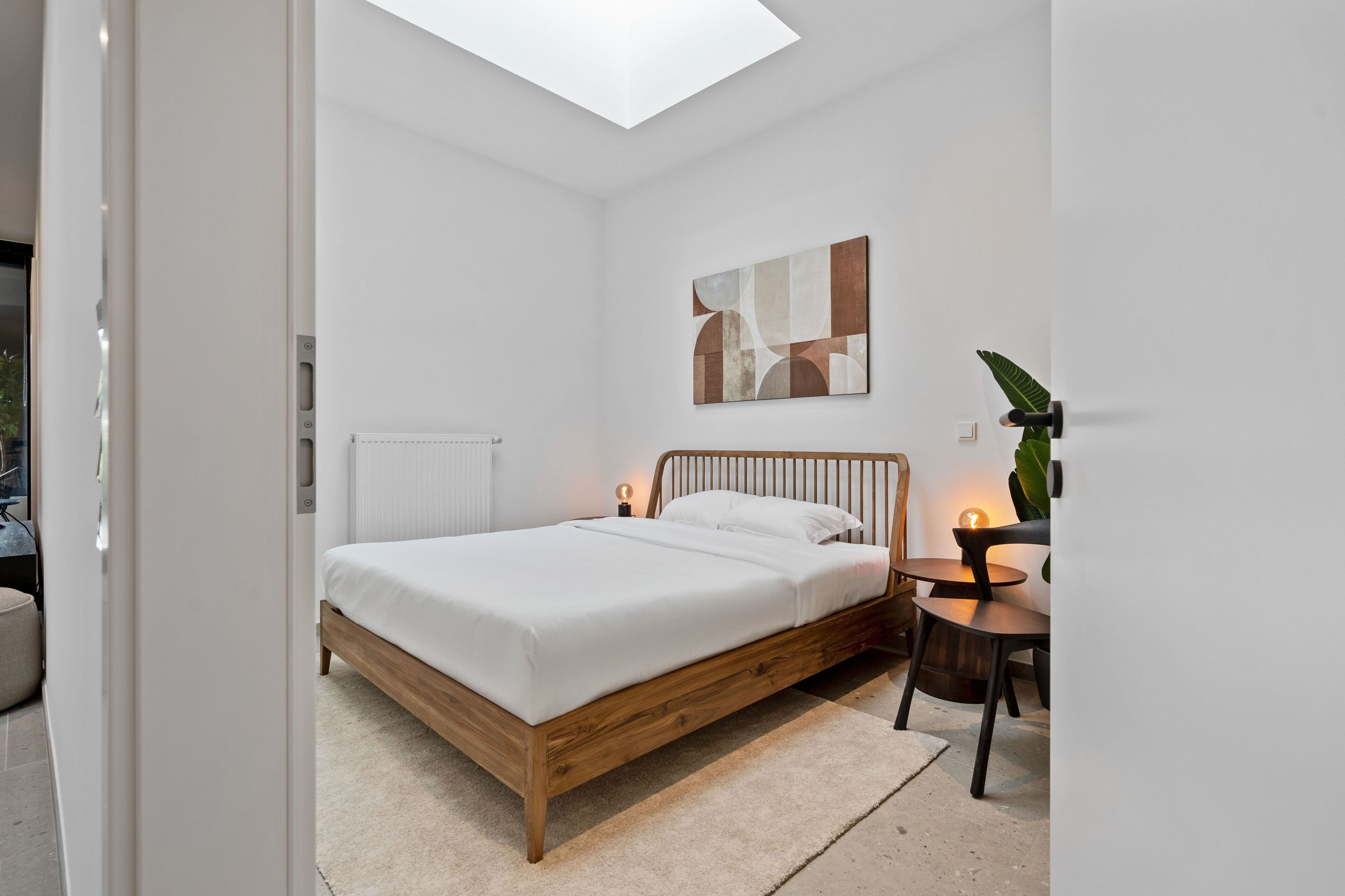 Een moderne slaapkamer met dakraam, een houten bed, nachtkastje met lamp en een abstract schilderij aan de muur.