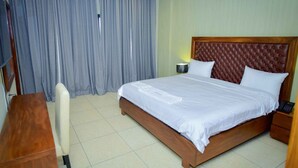 A hotel room with a large bed, a wooden desk, a chair, and a nightstand with a lamp. - Robyson Residency Kinshasa (Kinshasa)