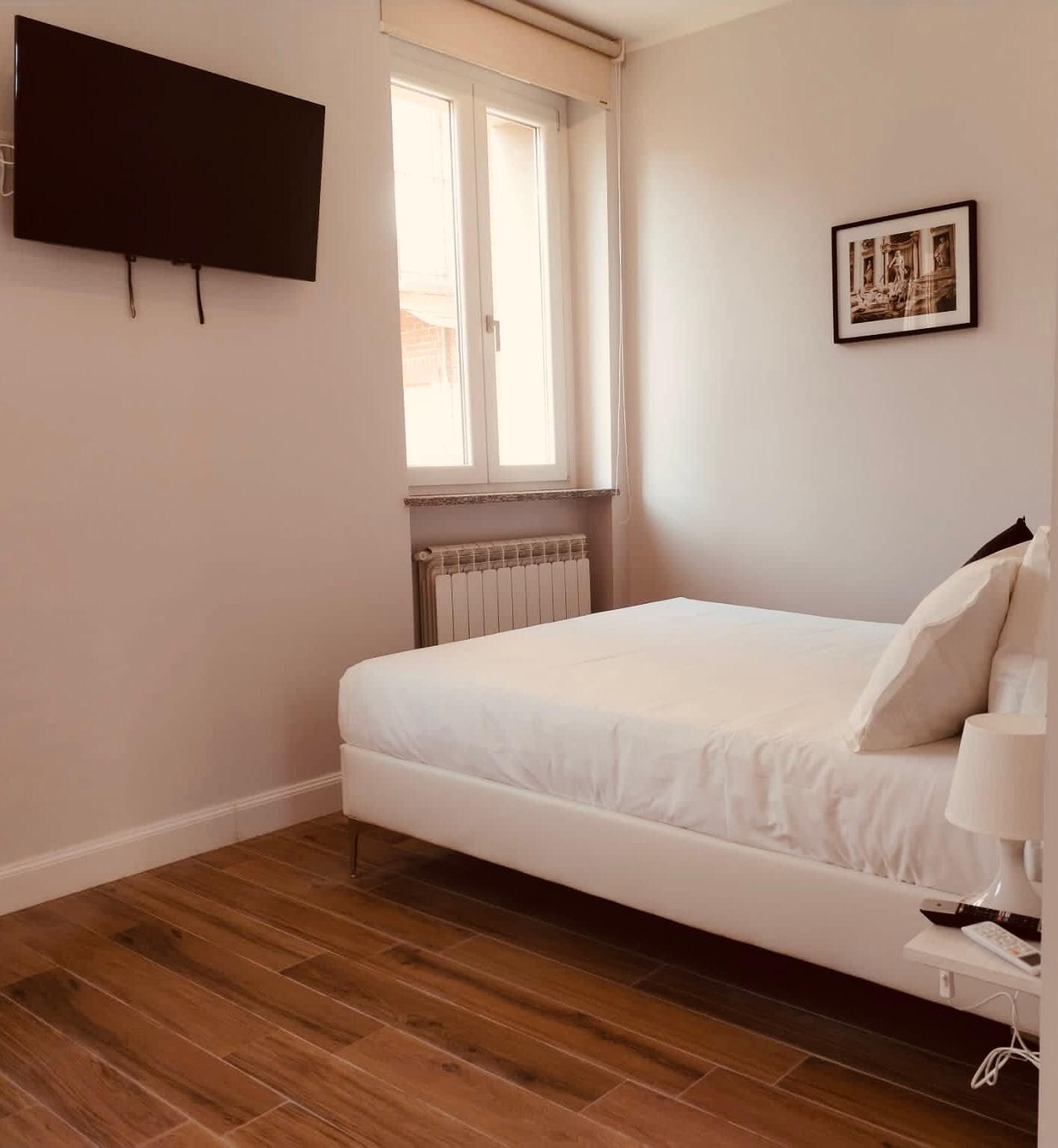 A bedroom with a bed, a window, a radiator, a TV mounted on the wall, and a framed picture.