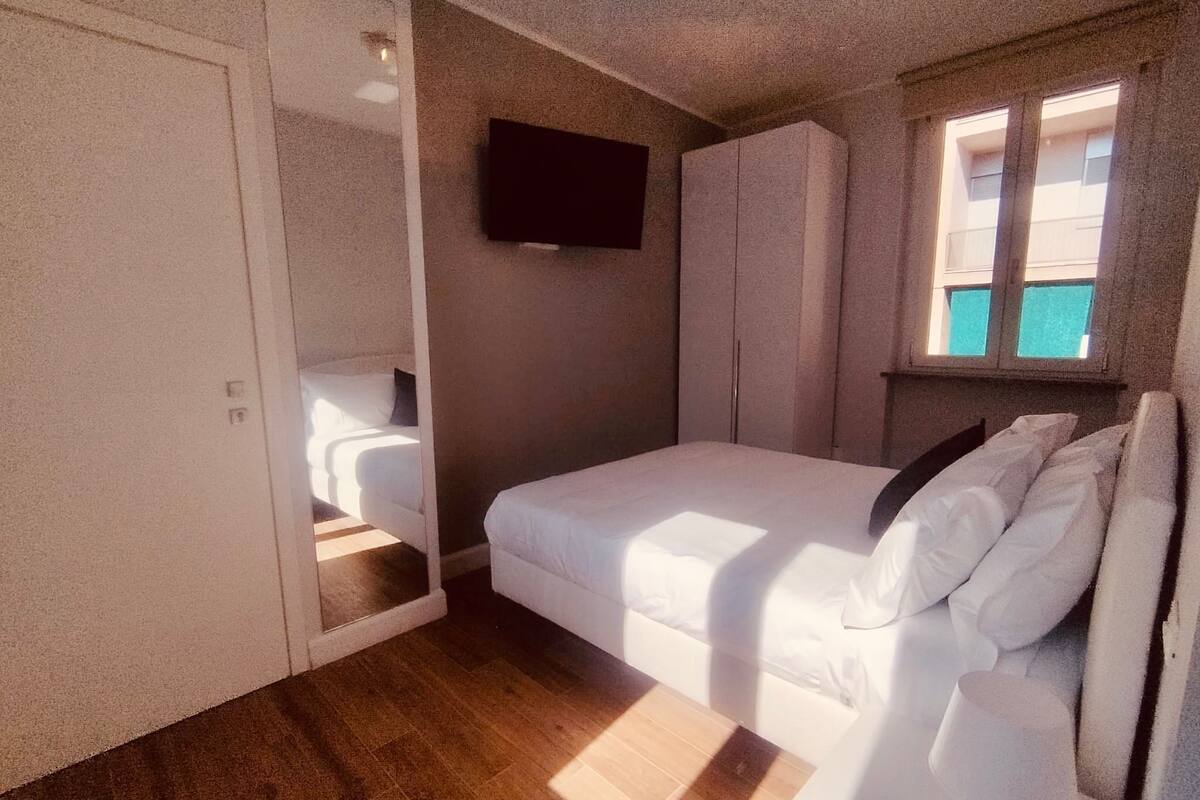 A bedroom with a bed, a TV mounted on the wall, and a window with a view of buildings.